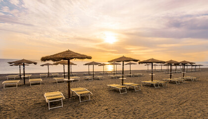  empty beach during beautiful sunrise or sunset with chaise loungues and nice umbrellas with blue sea, sun glow and amazing cloudy sky on thr background