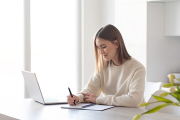 A young beautiful woman working on a laptop, sitting in the kitchen at home, remote work, freelance.