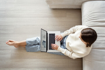 A young woman in casual clothes with a laptop is sitting on the floor in the living room. Shopping for clothes online in an online store.