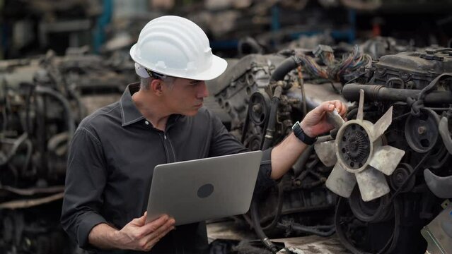 senior engineer using laptop computer checking list old used car engine in machinery parts shop warehouse. old technician man Repair vehicle automobile machine in Recycle motor factory