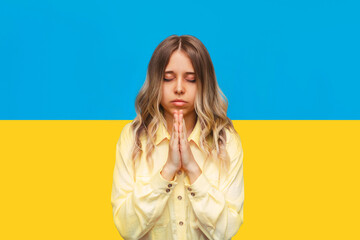 A young caucasian blonde woman prays with her eyes closed and folded hands asking help, rescue, peace isolated on a background of the Ukrainian flag