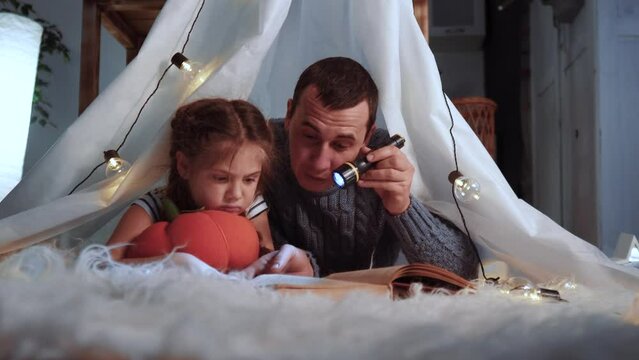 happy family at home dream. dad and daughter happy family at home lockdown reading a book in the evening in stay home homemade wigwam house. kid dream coronavirus concept. dad daughter read book