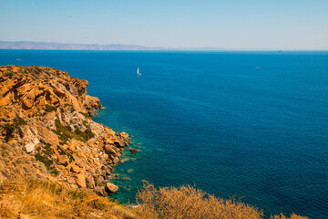 Fototapeta premium View from Cape Sounion (Greece) on Mediterranean Sea looking east with a sail boat in the background and the orange sand rocks in the foreground
