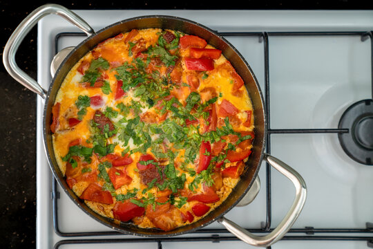 Scrambled eggs with vegetables and herbs in black frying pan on white stove. Top view
