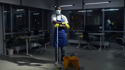 Full-length of African-American janitor in safety mask posing at camera in dark empty office