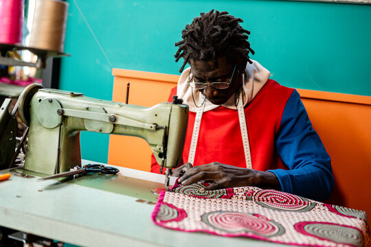 African adult tailor at work, artisan clothing manufacturer sewing with a vintage sewing machine, senegal wax traditional fabric, small business entrepreneur concept - Powered by Adobe