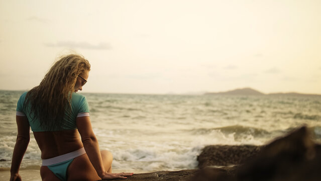 Silhouette sexy woman leaning on a rock reef enjoying and relax warm dark golden sunset. Back view. Woman in blue swimsuit and sunglasses. Concept rest tropical tourism happy summer holidays