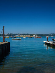 A view from a slipway across Bembridge Harbour, with yachts and boats floating on the mirror-like waters ahead of wooded hills and under blue skies.