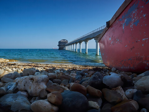 A low angle view from the shade of a derelict boat on the shore at Bembridge, including an expansive blue sky-and-sea, and the local lifeboat station.