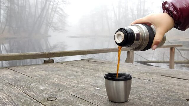 Woman pours tea from a thermos into a cup. Metal thermos with black tea. Outdoor recreation. Winter walk. Video 4k. Hot drinks outdoors.