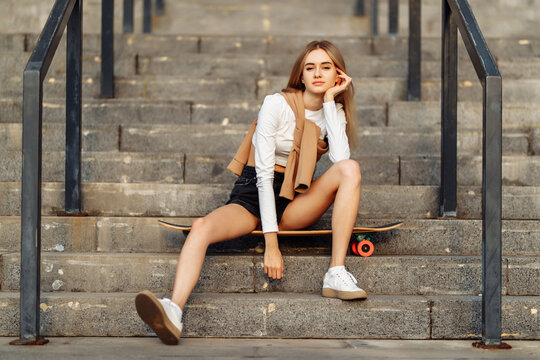 Cool young woman with a longboard sitting on the stairs in sunglasses. Casual style