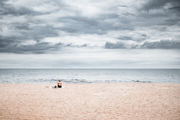 An woman and her dog sit alone on the beach watching a storm roll in.