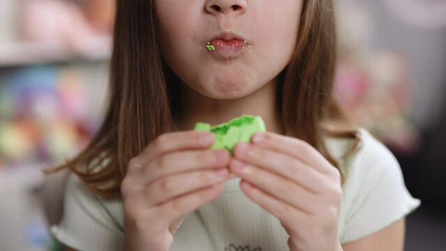 Close up of gorgeous school girl biting french macaroon, enjoying eating, confectionary desire and pleasure. Attractive brunette female girl on home background, licks her lips, eating macaron