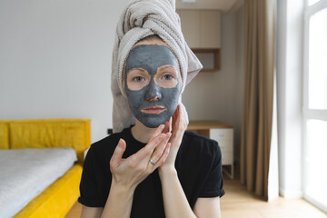 Young caucasian woman is applying greyish-blue mask on face sitting in her bedroom. Her hair is wrapped in a towel. Spa beauty procedures at home for girl. healthy skin care treatment concept.