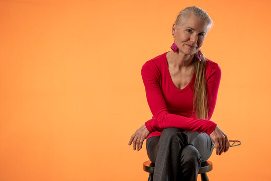 Beautiful Smiling Happy Blond Mature Woman On Solid Orange Background With Copy Space Sitting On Stool.