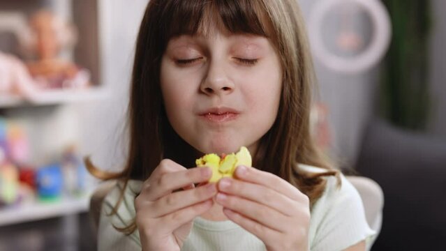 Attractive brunette school girl on a home background, licks her lips, eating one yellow macaron, closes her eyes with pleasure. Dessert person, sweet tooth, gourmet. Feeling good, delicious