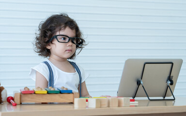 Little Caucasian nerd kid girl wearing glasses online learning on tablet at home and playing wooden block toys with colorful xylophone and puzzle pieces. White background
