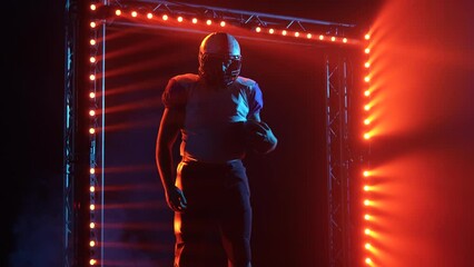 Unrecognizable confident quarterback holding ball. American football player in uniform and helmet stands on stadium in dark against red blue backlight and smoke. Waiting for start of match, set to win