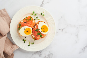 Sandwich with delicious toppings, smoked salmon, eggs, herbs and microgreens radish, black sesame seeds over white plate on white marble table background. Healthy open sandwich superfood. Top view.