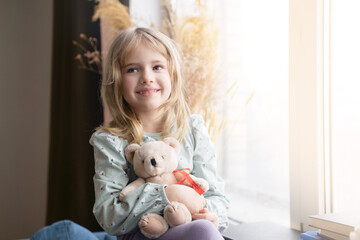 Сute  little kid girl hugging bear, sitting near window at home alone. Joyful child daughter holding toy, feeling happiness in kindergarten, looking aside.