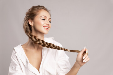Happy pretty young woman holding her long pigtail, pleased with healthy shining hairs posing on gray background. Beauty portrait of smiling girl with stylish braid. Haircare treatment products ad. - obrazy, fototapety, plakaty