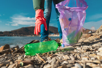 A volunteer in rubber gloves reaches for a dirty plastic bottle lying on the ocean shore. Hands close-up. Environmental pollution