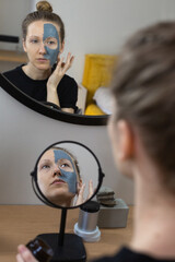rear view photo of a young caucasian woman looking at herself in small mirror, applying mud mask other face, reflection of beauty treatment in the big round mirror on the wall at home