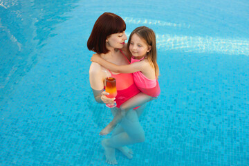 Fototapeta na wymiar Pretty young mom and daughter in pink swimsuits swimming in the pool with a drink in mother hand. Little cute beautiful girl hugs mom in an exotic pool with turquoise water.