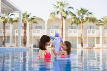 Fototapeta na wymiar Little girl with her mom having fun in swimming pool. A young mother teaching her daughter swim in clear blue water in a pool with an inflatable lifebuoy in the form of a unicorn.
