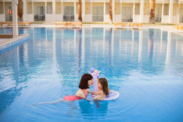 Young girl kid with unicorn inflatable ring splashing in swimming pool having fun leisure activity. Happy mother and her daughter in the same pink swimsuits playing in swimming pool. Summer vacations