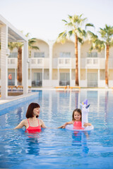 Charming young red-haired mom in a pink bikini swimsuit holding hand of her beautiful little daughter in a lifebuoy while teaching her to swim in the pool during a relaxing time at a luxury resort