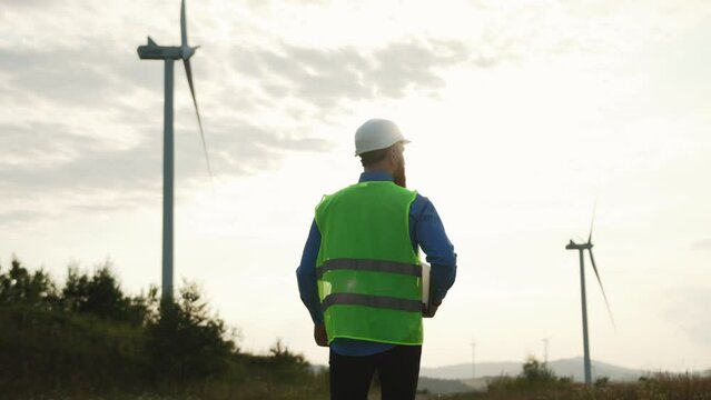 Caucasian technician worker in green safety vest walking along operative wind turbines. Chief engineer of windmill farm working on industrial factory. Ecology concept.