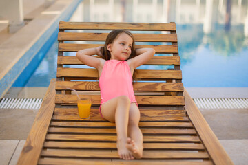 A little girl with blond hair putting hands behind head relaxing sunbathing in a pink swimsuit. The child is on vacation, siting on a sun lounger. Bright summer photo.