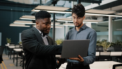 Two male business partners men Arabian man holding laptop showing presentation African businessman looking at computer screen colleagues working online discuss startup pointing display making choice