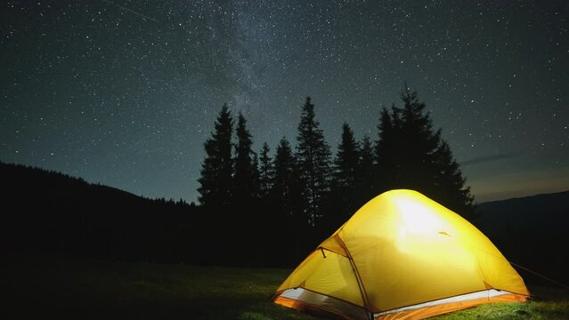 Time lapse of brightly illuminated camping tent glowing on campsite in dark mountains under night stars covered sky. Active lifestyle and traveling concept