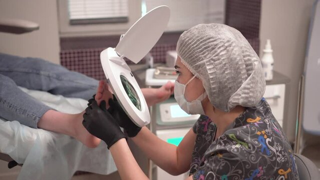 The pedicure master looks at the patient's foot through a large magnifying glass. Procedure at the podiatrist in the salon.