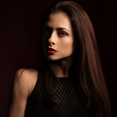 Fototapeta premium Beautiful bright makeup woman with red lipstick and long brown healthy hair with half face in shadow on dark red shadow background. Closeup