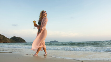 Fototapeta na wymiar Sexy hot woman walks on golden sunset on tropical beach, wet sand, against background of sea. Girl in green swimsuit and pink tunic silk shirt cape, drinks her orange cocktail Pina Colada.