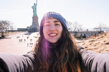 Young woman taking a selfie smiling with the statue of liberty in the background while sightseeing....