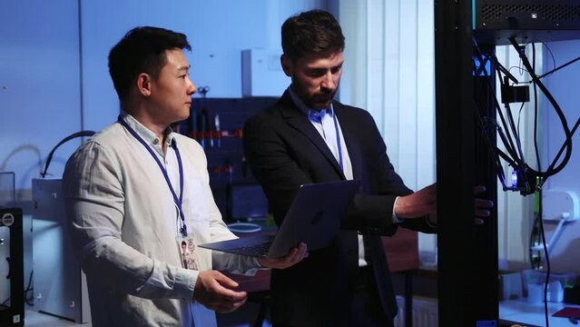 Asian lab engineer cooperating with caucasian employee explaining work of 3D dimensional printer. Laboratory teamwork. Electric engineering. Cooperation.
