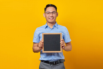 Portrait of smiling young Asian businessman in glasses holding chalkboard and standing confident looking at camera isolated over yellow background