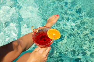 Female model is holding glass of cocktail by the blue pool. glass of colorful fruit cocktail