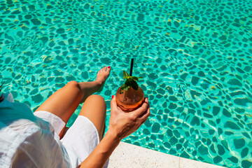 Female model is holding glass of cocktail by the blue pool. glass of colorful fruit cocktail