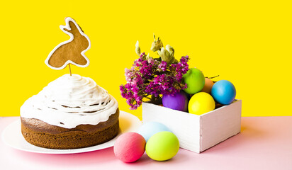 On a yellow background, an Easter cake decorated with cookies in the shape of a rabbit in a wooden box, multi-colored eggs and delicate flowers.  Easter bright holiday concept.  Front view.