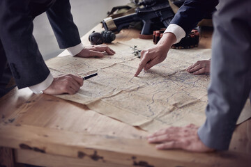 Planning a hostile take-over. Two men in suits discussing plans while guns and a map lie on a table.