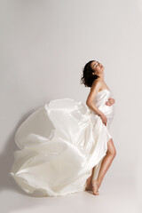 Happy woman waiting for a baby, wrapped in flowing white silk on a white background. The concept of...