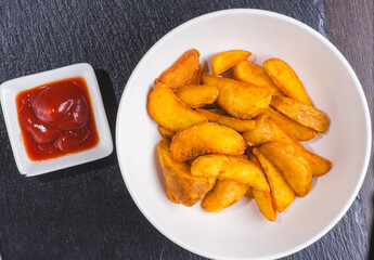 Rustic potatoes with ketchup on light wooden plate on dark wooden background.