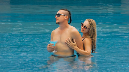 Obraz na płótnie Canvas The loving couple hugs and kisses, drinking blue cocktail alcohol liquor in swimming pool at hotel outdoor. Portrait of caucasian man and woman. Creative hairstyles bodybuilder, swimsuit, sunglasses.