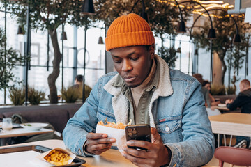 Positive African-American guy in orange hat eats tasty hamburger looking into mobile phone at table...