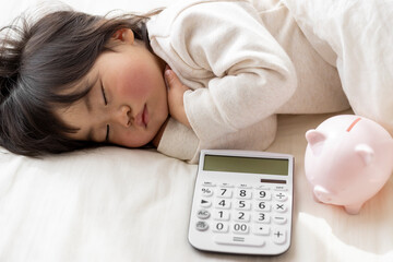 昼寝している赤ちゃんと電卓と豚の貯金箱(1歳8ヶ月、日本人、女の子)。育児の費用イメージ